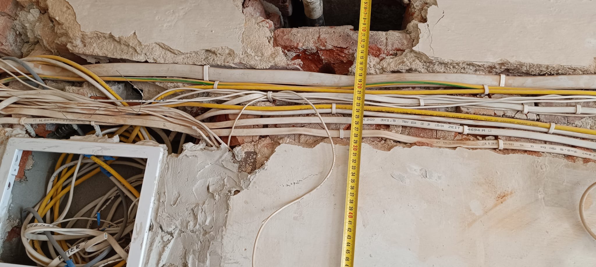 Odsłonięte przewody elektryczne w ścianie podczas remontu, częściowo zamurowane, z widoczną miarką. Żółte i białe kable, fragment puszki elektrycznej.