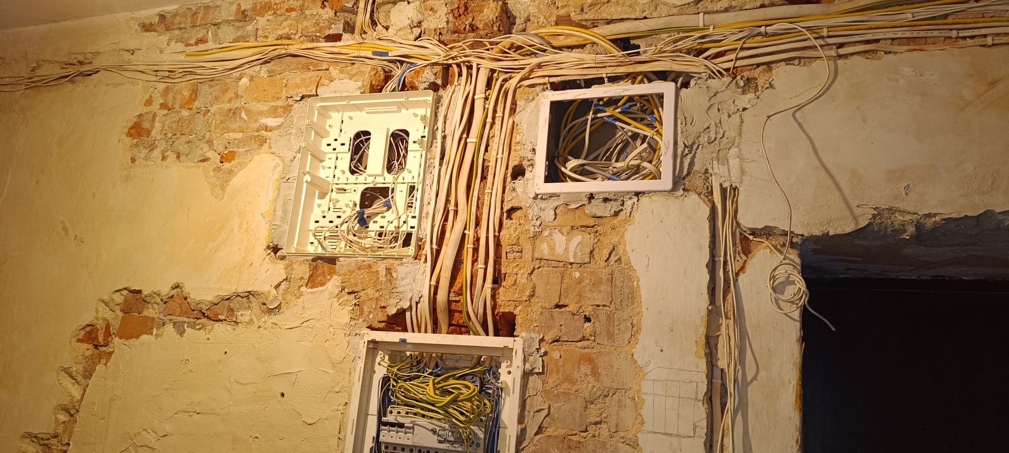 Ściana z odsłoniętą cegłą, na której zamontowano trzy puste skrzynki elektryczne z wystającymi kablami. Widoczne przewody w żółtej i białej izolacji.
