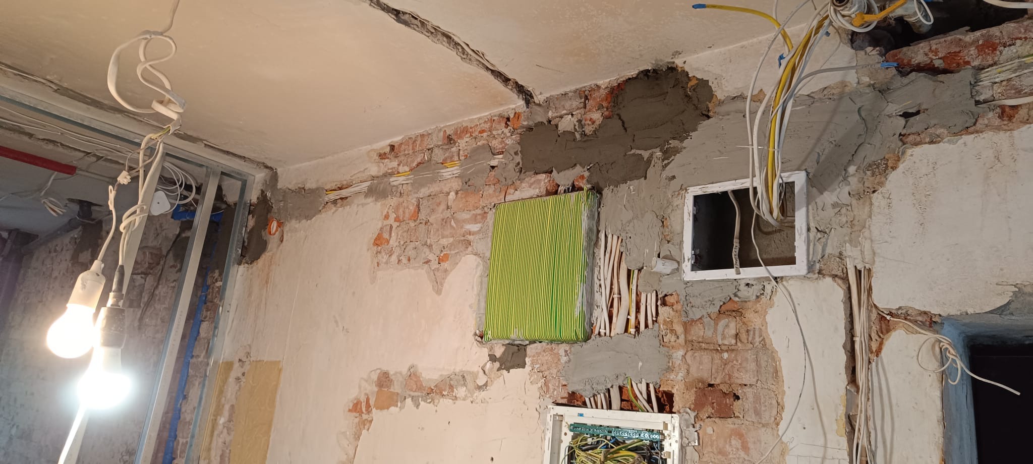 Remont pokoju: odsłonięta cegła, świeże tynki, instalacja elektryczna w ścianie, wiszące kable, puszki elektryczne. W tle metalowa konstrukcja ścianki działowej.