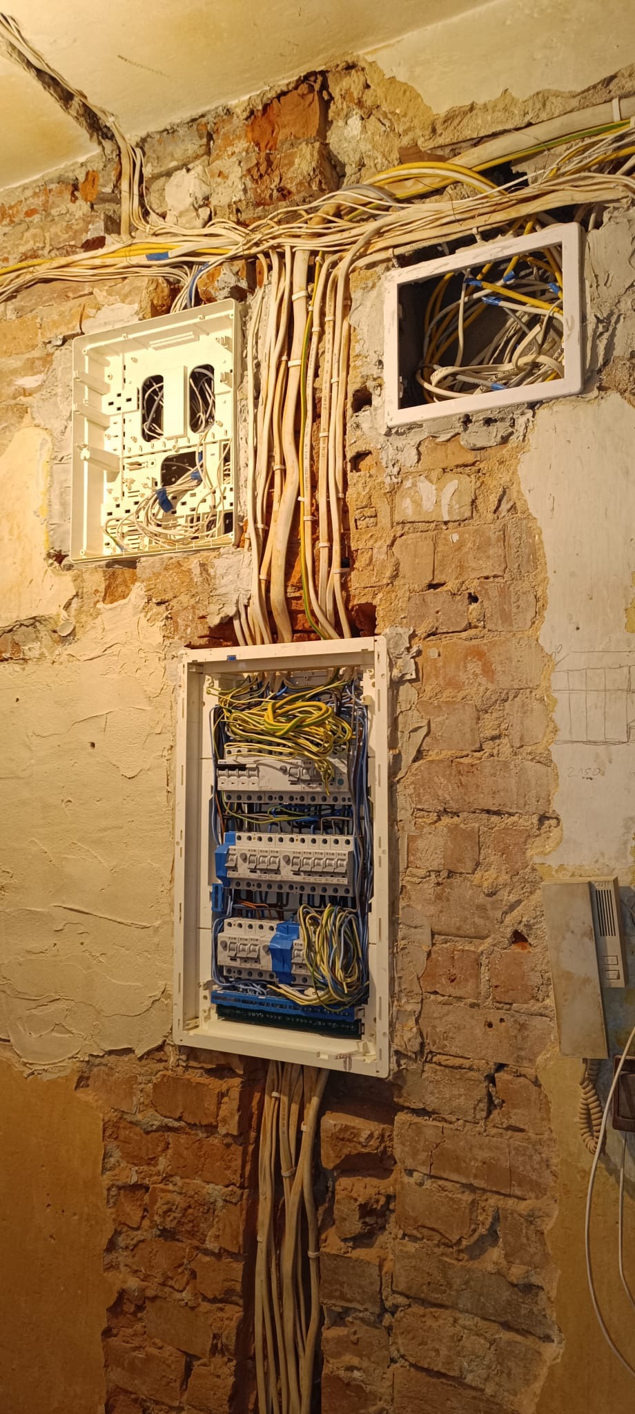 Rozdzielnia elektryczna w trakcie montażu na ceglanej ścianie. Widoczne kable i puszki instalacyjne. Częściowo otynkowana ściana.
