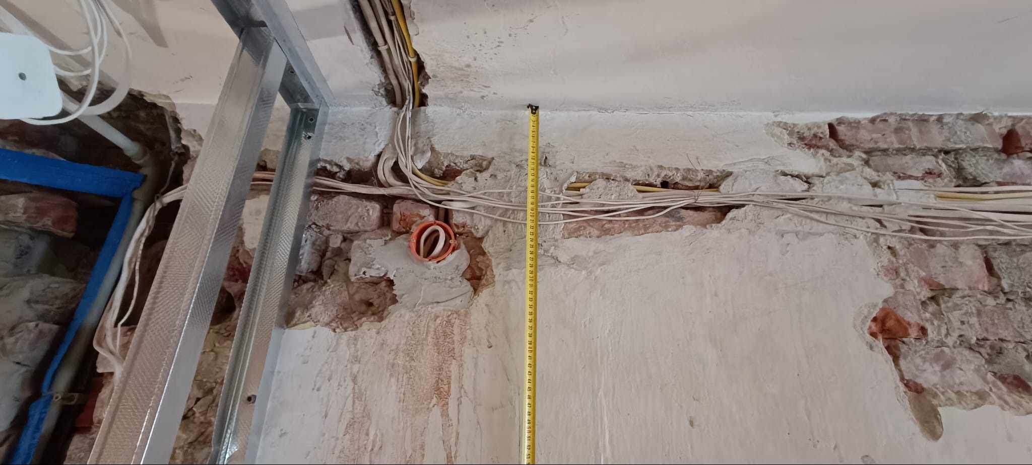 Ściana z odsłoniętą cegłą i instalacją elektryczną, częściowo zakryta płytą gipsową. Widoczna pionowa miarka, przewody i puszka elektryczna. Prace remontowe w toku.