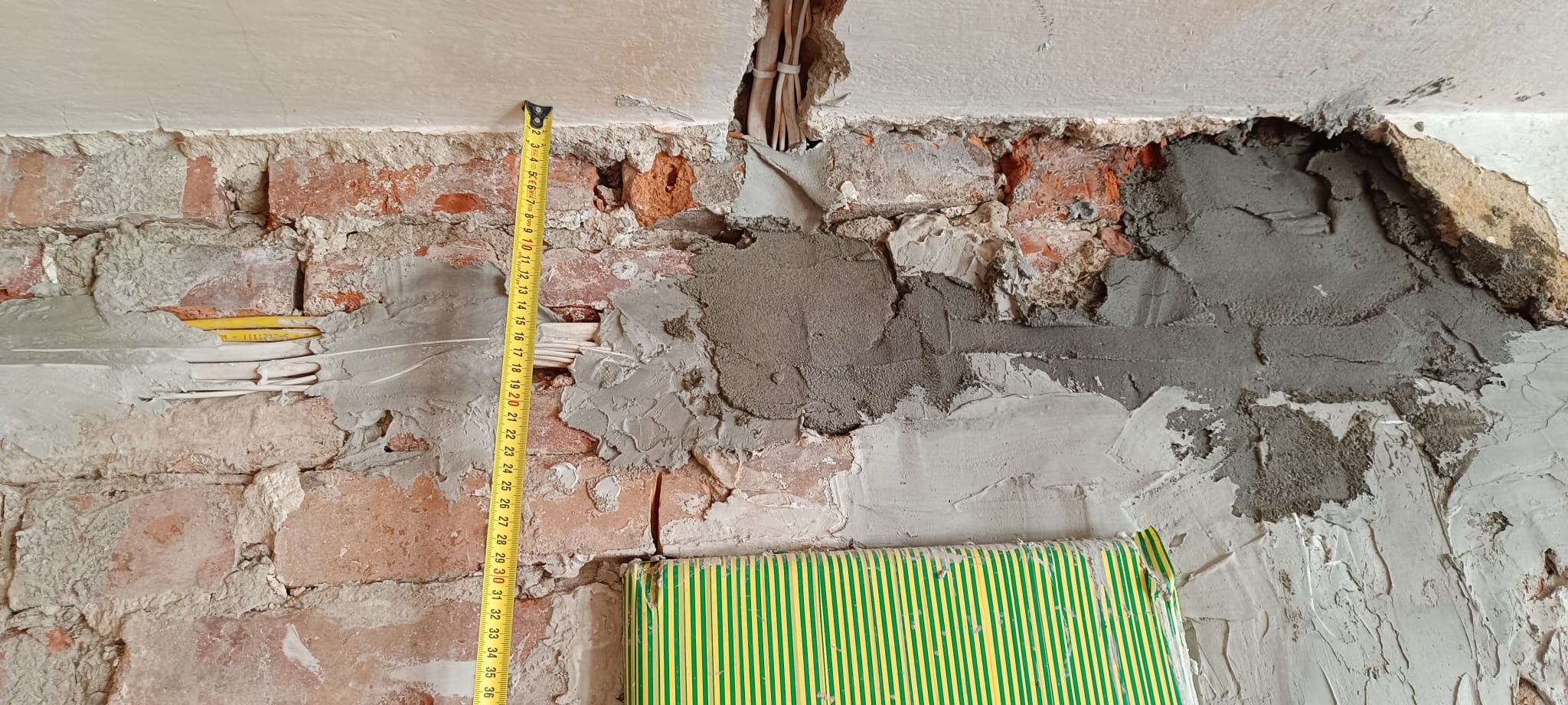 Nierówna ściana z cegły po kuciu, częściowo pokryta zaprawą. Widoczne kable elektryczne i miara zwinięta. Prace remontowe w trakcie, surowy stan.