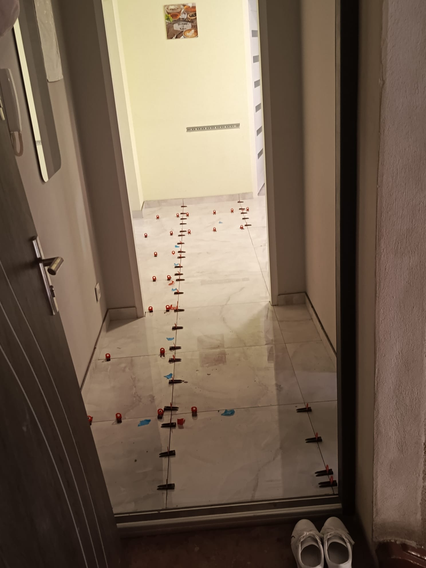 Układanie jasnych płytek podłogowych w korytarzu z użyciem systemu poziomowania, widoczne krzyżyki dystansowe i kliny. W tle obrazek z motywem kawy.