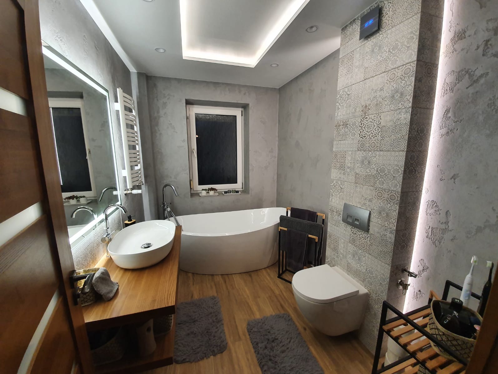 Nowoczesna łazienka z owalną wanną, drewnianym blatem pod umywalką i szarą mozaiką na ścianie. Oświetlenie LED podkreśla elegancki wystrój.