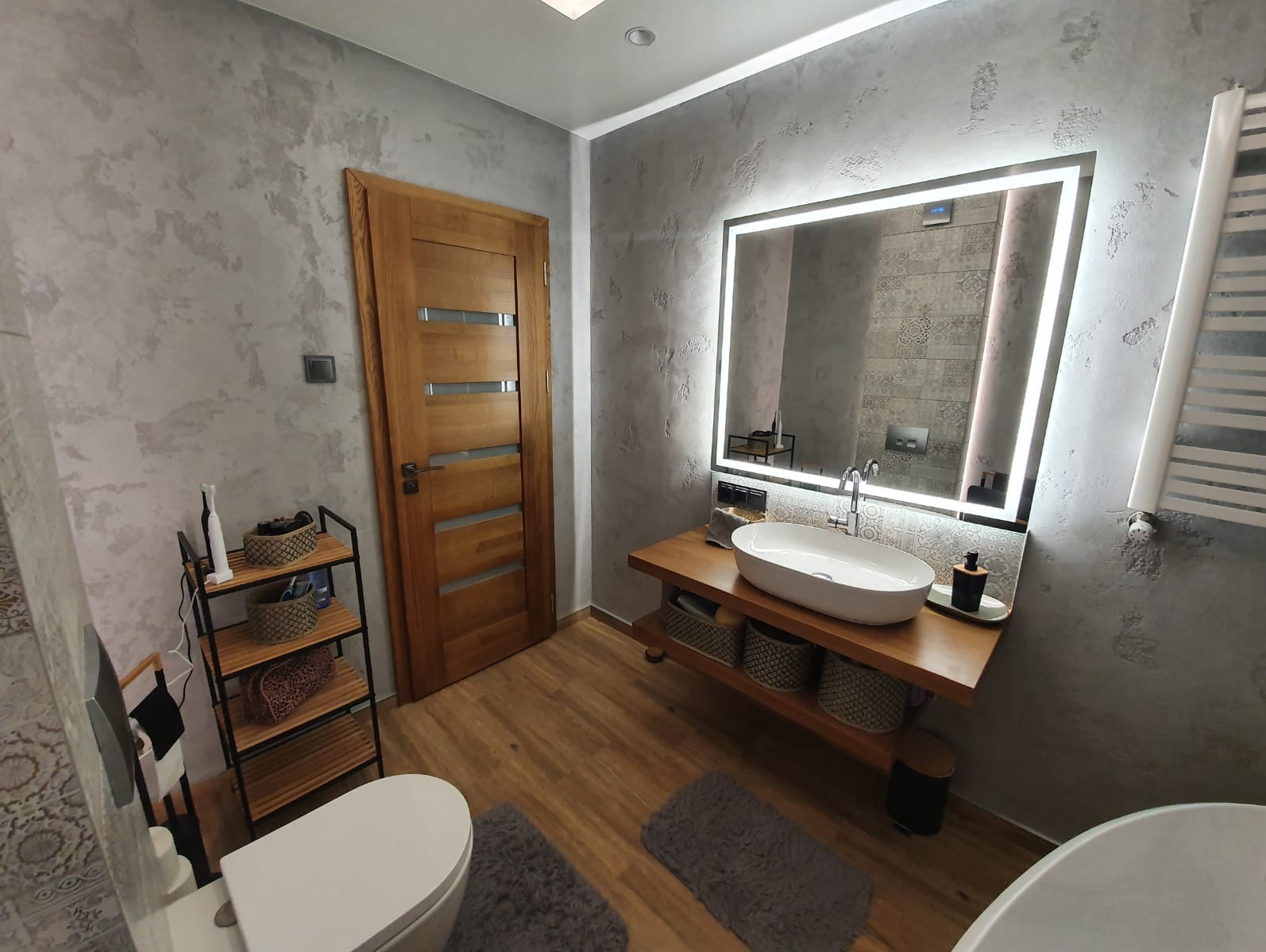 Stylowa łazienka z drewnianymi akcentami: dębowe drzwi, umywalka na blacie z półkami, lustro z podświetleniem LED i dekoracyjna ściana w odcieniach szarości.