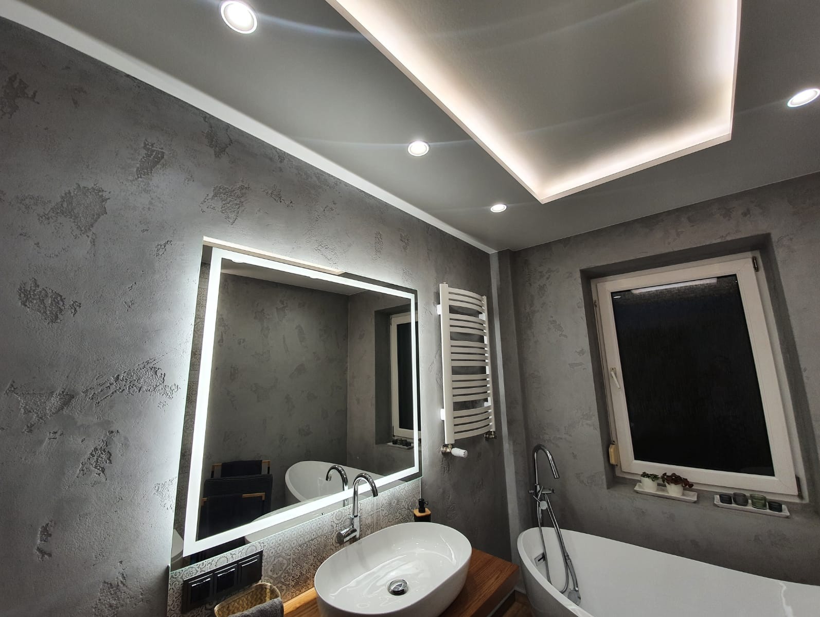 Szara łazienka z oświetleniem LED: lustro z ramą świetlną, grzejnik drabinkowy i owalna umywalka. Ściany w fakturze betonu, sufit z podświetleniem.