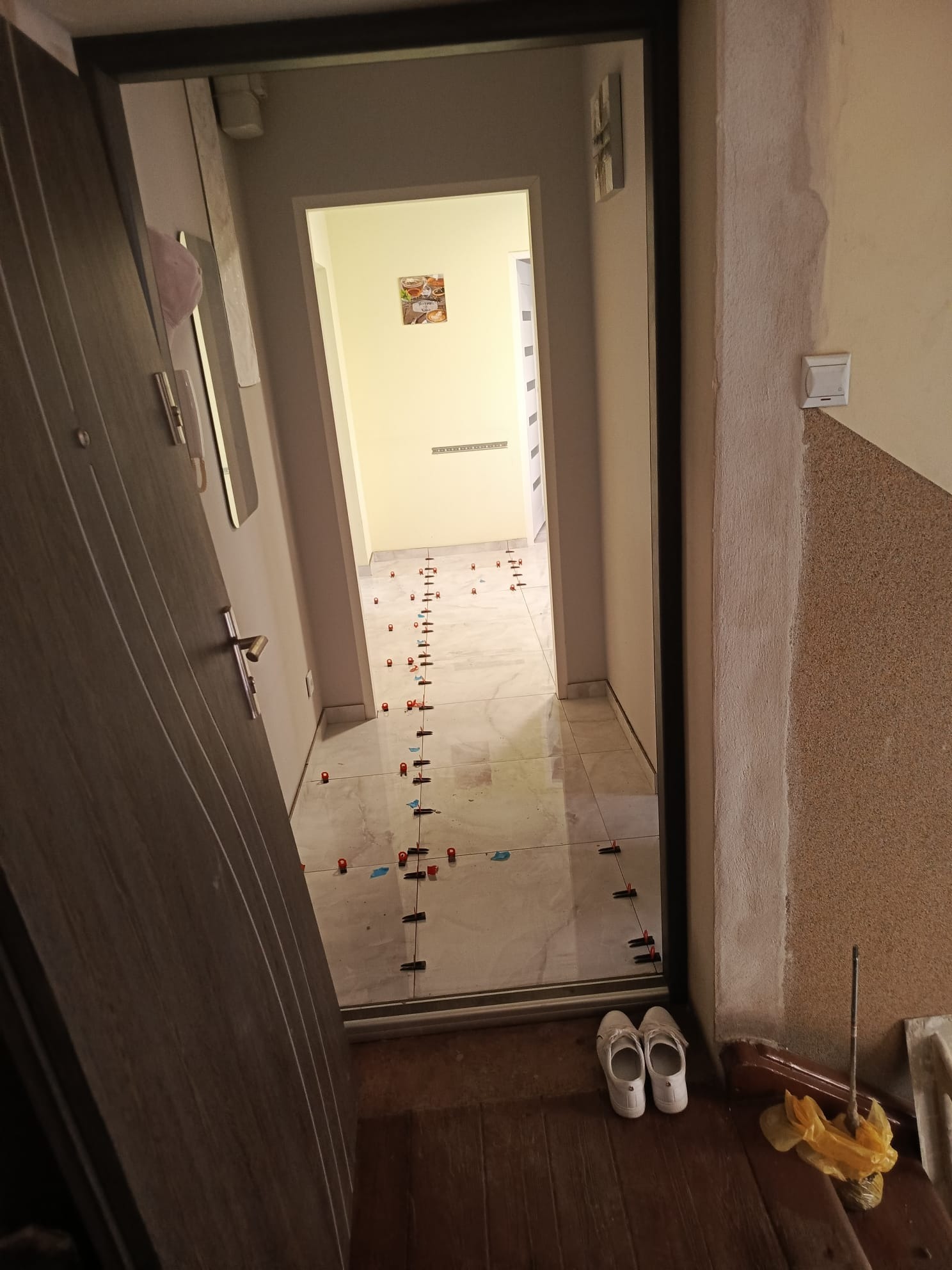 Korytarz z jasnymi płytkami podłogowymi, w trakcie układania, widoczne krzyżyki dystansowe. Otwarte drzwi wejściowe z lewej strony, w tle dalsza część mieszkania.