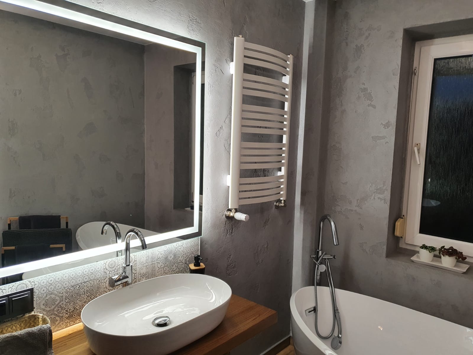 Nowoczesna łazienka w szarościach: owalna umywalka na drewnianym blacie, podświetlane lustro, grzejnik drabinkowy i wanna. Dekoracyjna struktura ściany.