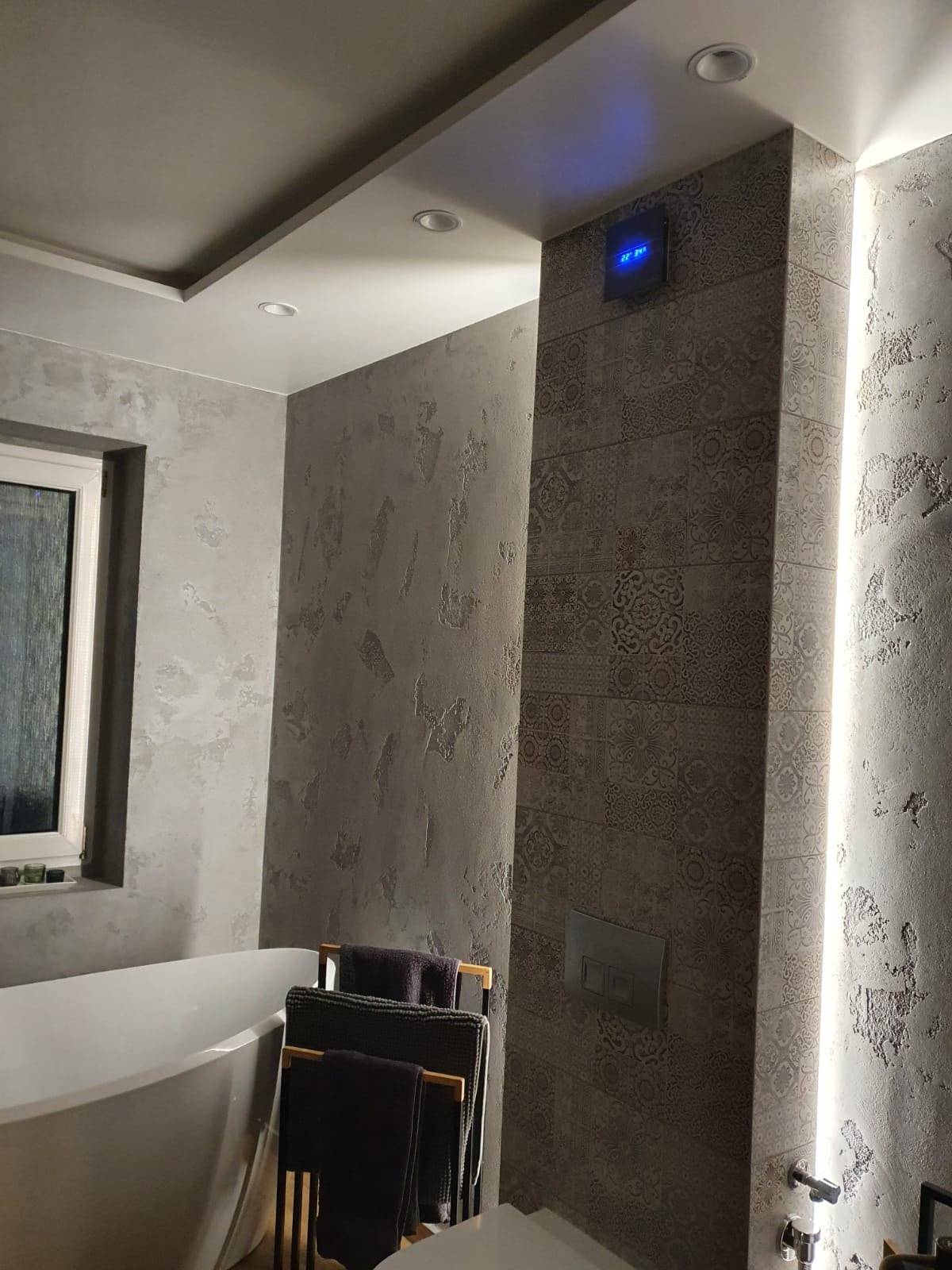 Wykończona łazienka z wanną, dekoracyjną ścianą z płytek wzorzystych i oświetleniem LED. Na ścianach szara struktura, a na suficie punktowe oświetlenie.