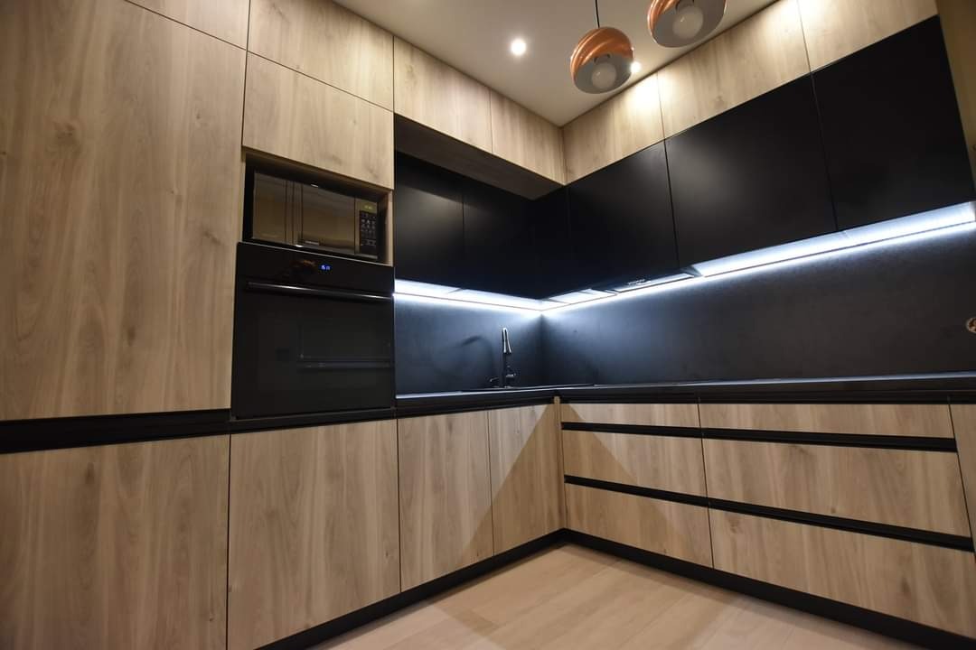 Nowoczesna kuchnia na wymiar z drewnianymi frontami i czarnymi elementami, oświetleniem LED pod szafkami. Widoczne AGD w zabudowie i minimalistyczne lampy.