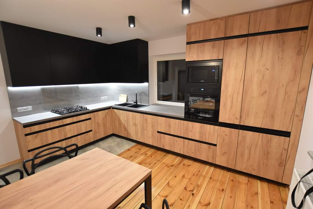 Nowoczesna kuchnia z drewnianymi frontami i czarnymi akcentami, oświetleniem LED pod szafkami i drewnianą podłogą. Widoczny stół z krzesłami.