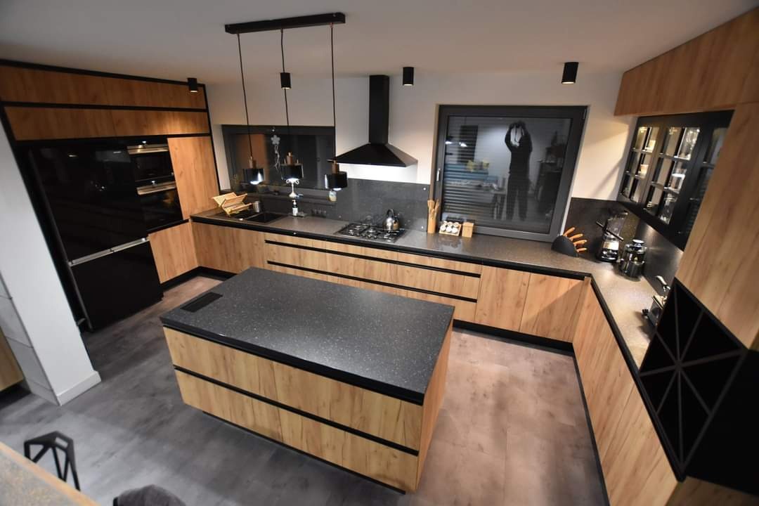 Nowoczesna kuchnia z wyspą, fronty w odcieniach drewna z czarnymi akcentami. Widoczne oświetlenie punktowe i okno z odbiciem osoby robiącej zdjęcie.