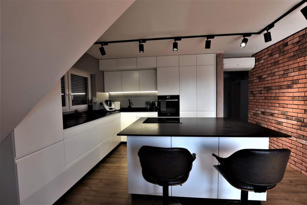 Nowoczesna kuchnia na poddaszu z białą zabudową, czarnym blatem i wyspą. Ceglana ściana i oświetlenie punktowe dodają charakteru. Dwa krzesła barowe przy wyspie.