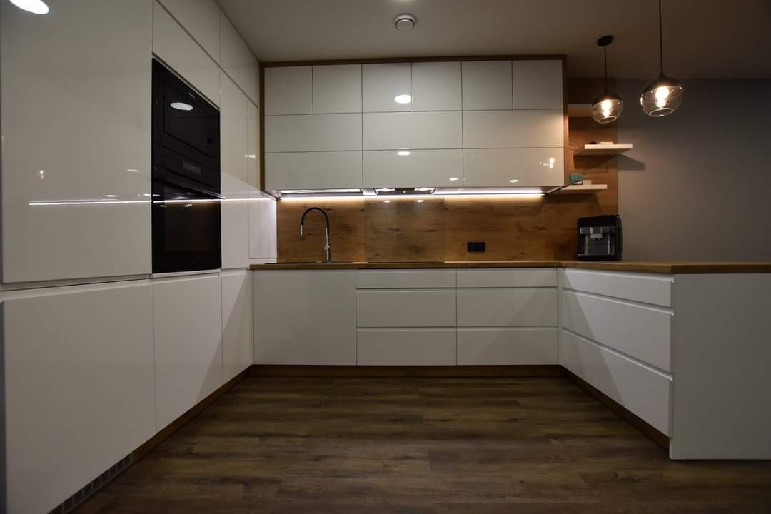 Nowoczesna kuchnia z białymi, lakierowanymi frontami, drewnianym blatem i oświetleniem LED pod szafkami. Widoczne wiszące lampy i ekspres do kawy.