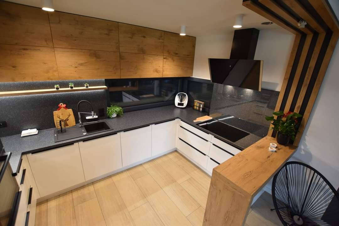 Nowoczesna kuchnia narożna z białymi szafkami, drewnianymi frontami górnymi i czarnym blatem. Uwagę zwraca dekoracyjny element z drewna i czarnych listew.