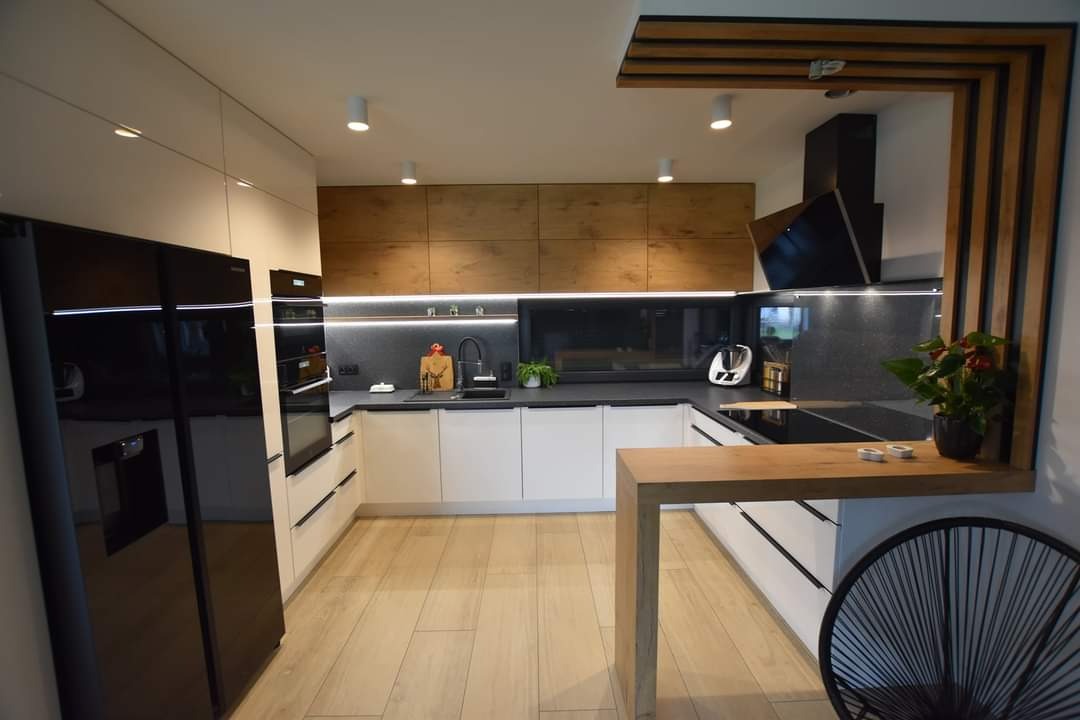 Nowoczesna kuchnia w kształcie litery U z białymi szafkami dolnymi i drewnianymi górnymi, czarnym blatem i sprzętem AGD. Widoczny fragment drewnianej konstrukcji nad blatem.