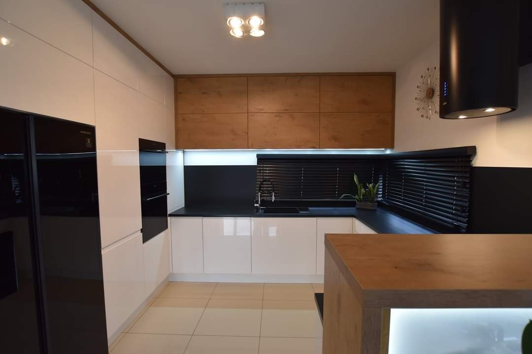 Nowoczesna kuchnia z białymi, lakierowanymi frontami, drewnianymi szafkami górnymi i czarnymi akcentami. Widoczny sprzęt AGD w zabudowie i rolety okienne.