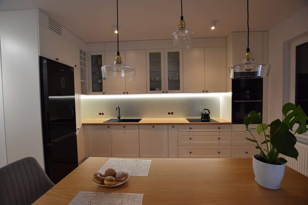 Jasna kuchnia na wymiar z drewnianym blatem, oświetleniem LED i szklanymi lampami. Na stole talerz z pączkami i roślina doniczkowa. Nowoczesny design.