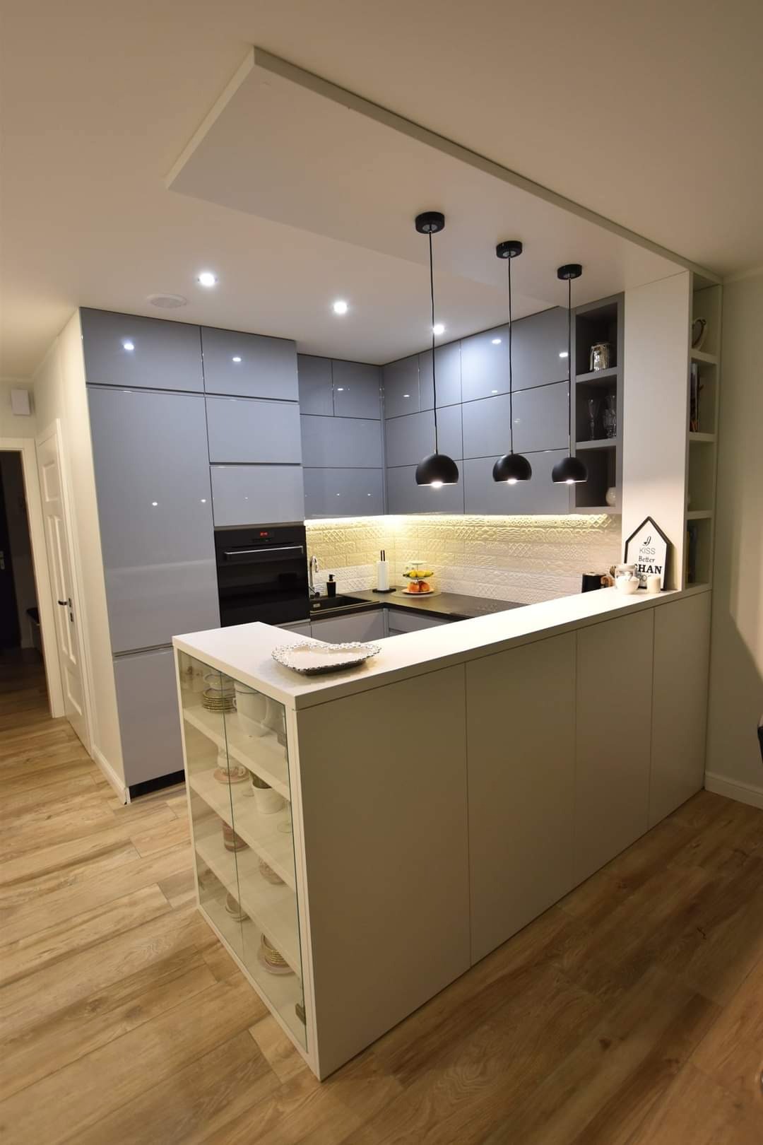 Nowoczesna kuchnia z szarymi, lakierowanymi frontami, oświetleniem punktowym i trzema czarnymi lampami wiszącymi nad blatem. Biała wyspa z przeszklonymi półkami.