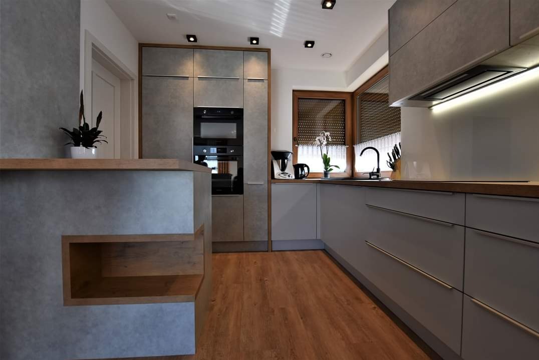 Nowoczesna kuchnia z szarymi frontami i drewnianymi akcentami. Widoczna zabudowa z piekarnikiem i mikrofalówką, wyspa z półkami i drewniana podłoga.