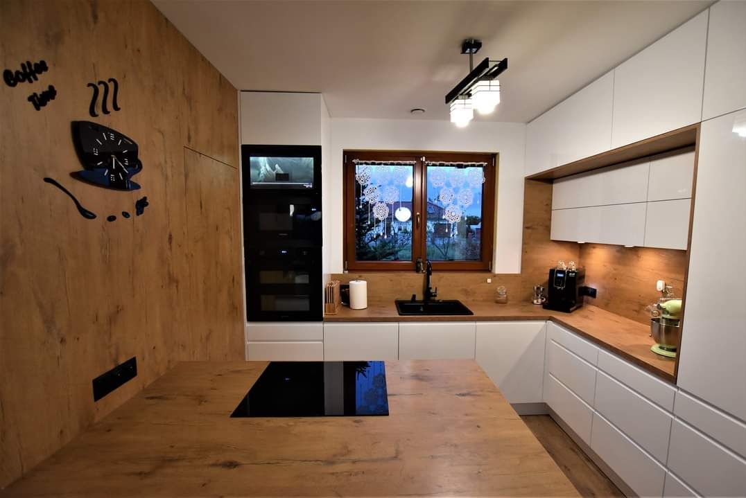 Nowoczesna kuchnia z białymi szafkami i drewnianymi blatami, czarnym zlewem i płytą indukcyjną. Na ścianie dekoracja 'Coffee Time' i okno z ozdobami.