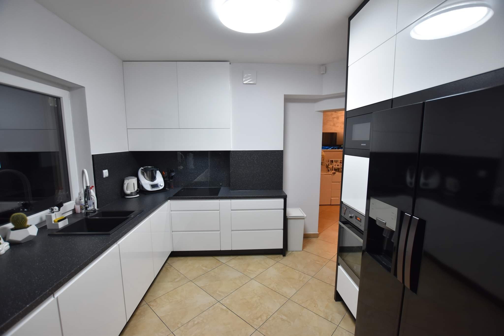 Nowoczesna kuchnia z białymi szafkami i czarnym blatem. Widoczny zlew, płyta indukcyjna, ekspres do kawy i lodówka w zabudowie. Jasne płytki na podłodze.