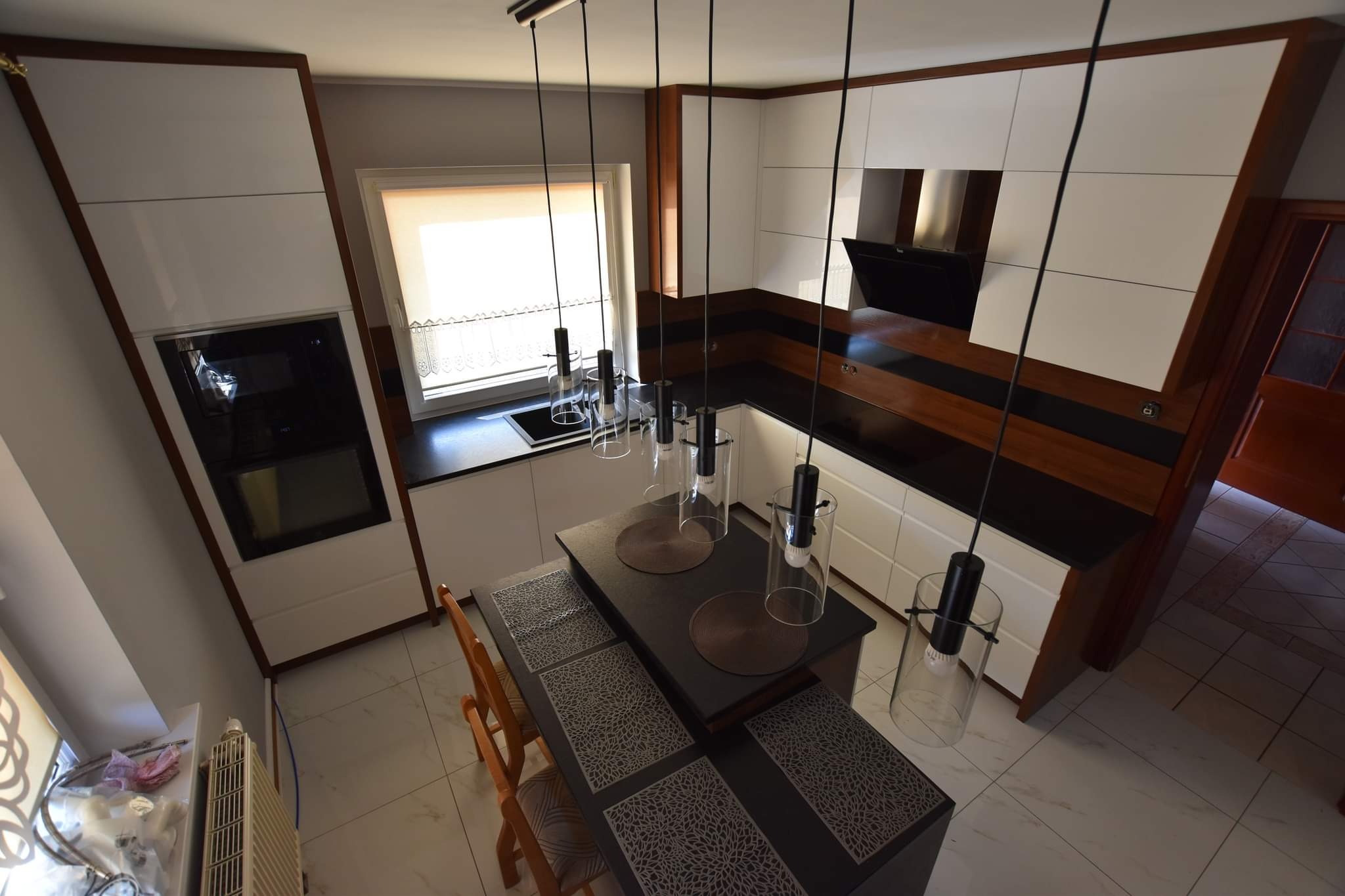 Nowoczesna kuchnia z białymi szafkami, drewnianymi akcentami i czarnym blatem, oświetlona wiszącymi lampami. Widok z góry na wyspę kuchenną z krzesłami.