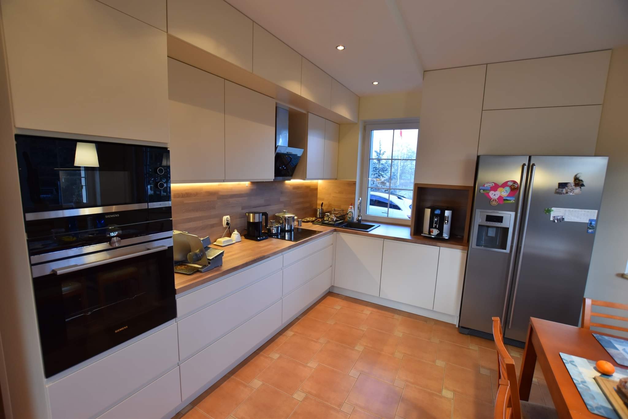 Jasna kuchnia z drewnianym blatem i nowoczesnym sprzętem Siemens, oświetlona listwą LED pod szafkami. Widoczna lodówka i stół z krzesłami.