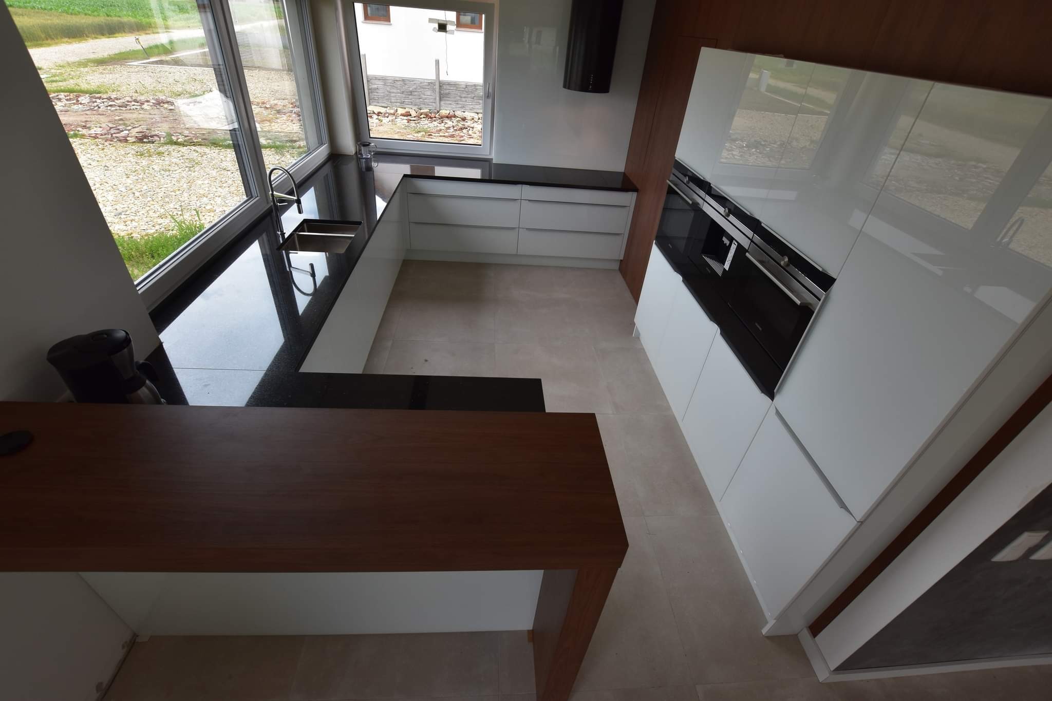 Nowoczesna kuchnia z białymi, lakierowanymi frontami, czarnym blatem i drewnianym barkiem. Duże okno z widokiem na zewnątrz. Minimalistyczny design.