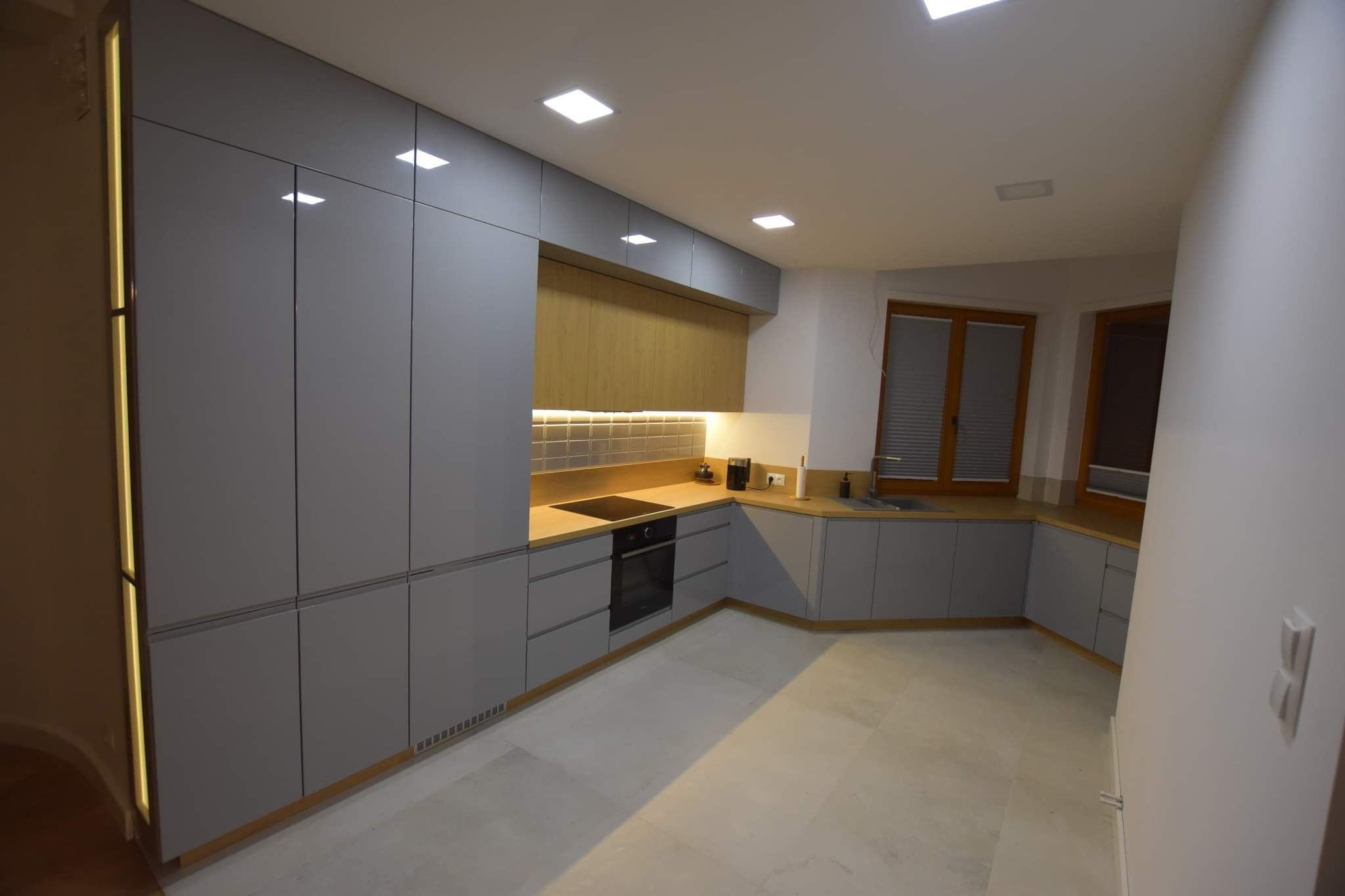 Nowoczesna kuchnia w odcieniach szarości z drewnianymi akcentami, oświetleniem LED i minimalistycznym designem. Widok na okno z żaluzjami.