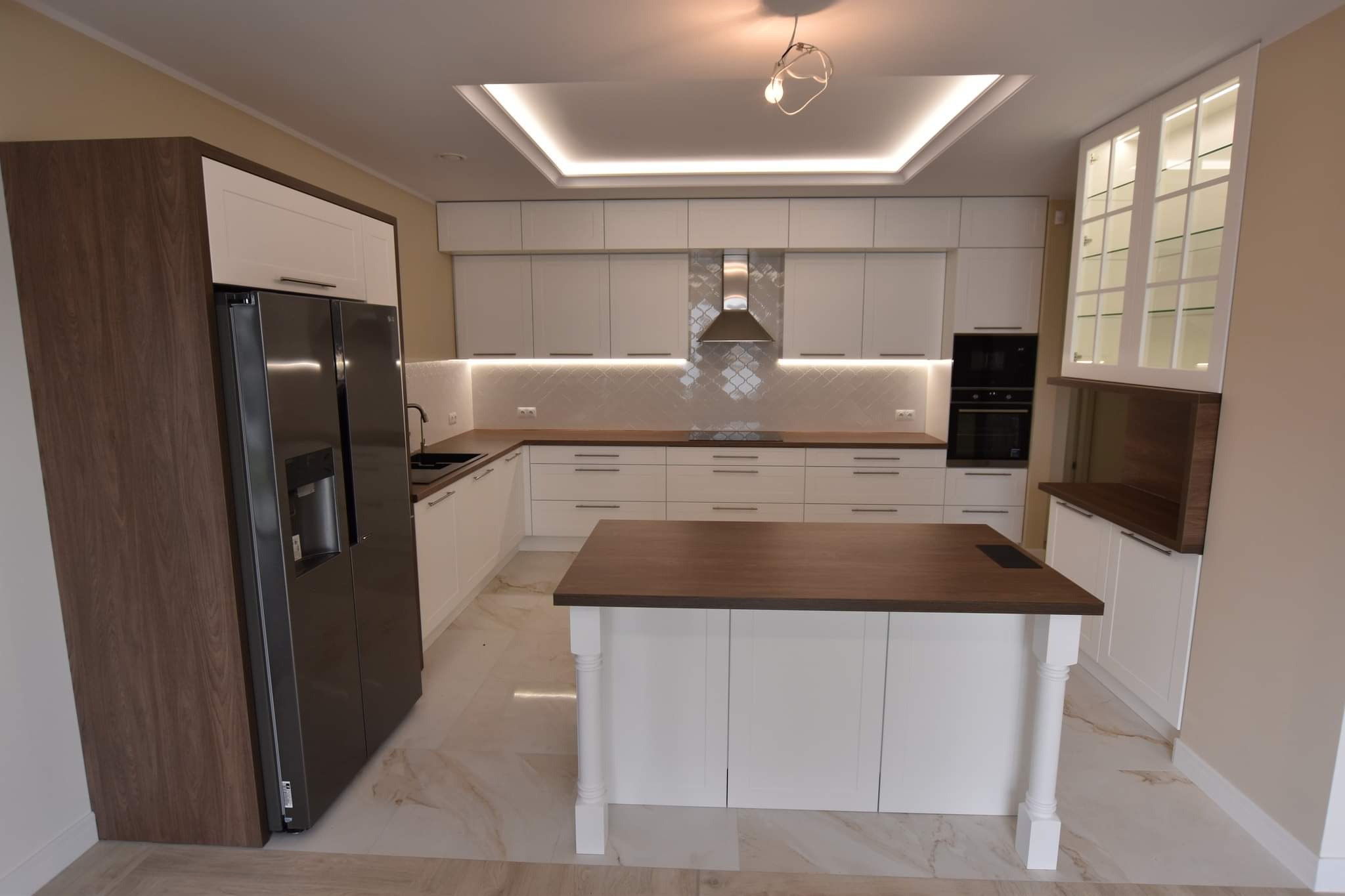 Nowoczesna kuchnia z wyspą, białe fronty szafek, drewniany blat, oświetlenie LED podszafkowe i sufitowe, duża lodówka z drewnianym wykończeniem.