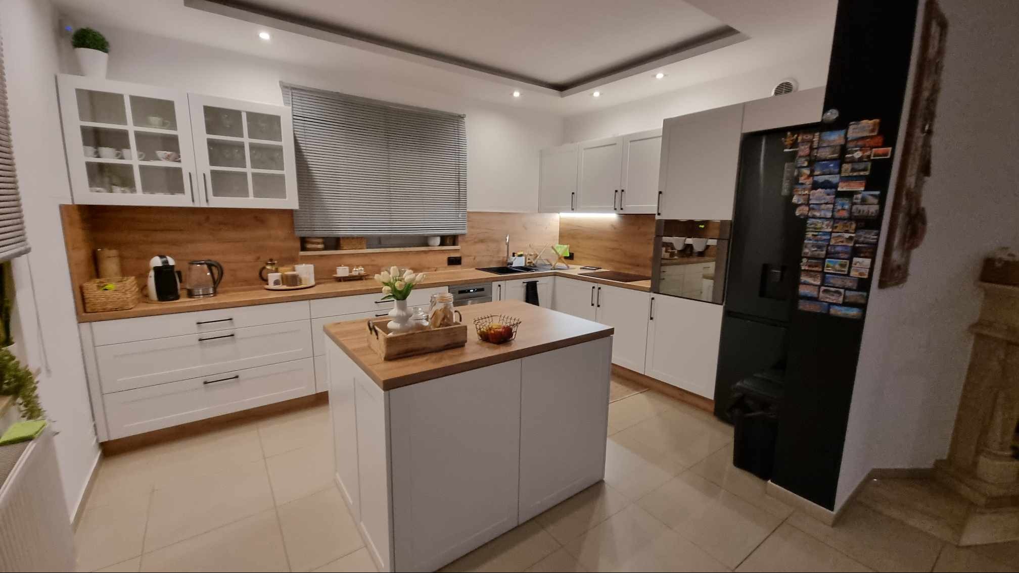 Nowoczesna kuchnia z wyspą, białe szafki z drewnianymi blatami, czarna lodówka z magnesami, rolety w oknie, oświetlenie sufitowe. Przytulne i funkcjonalne wnętrze.