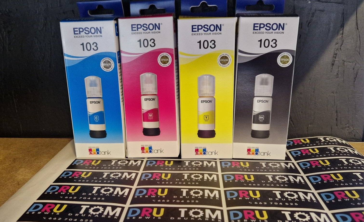 Cztery opakowania tuszy Epson 103 (C, M, Y, BK) na tle arkusza naklejek z logo 'DRU TOM Serwis Drukarek', ukazujące akcesoria do drukarek.
