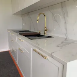 Biała, nowoczesna kuchnia z marmurowym blatem i złotą armaturą. Widoczny zlew, płyta grzewcza i eleganckie uchwyty. Minimalistyczny design.