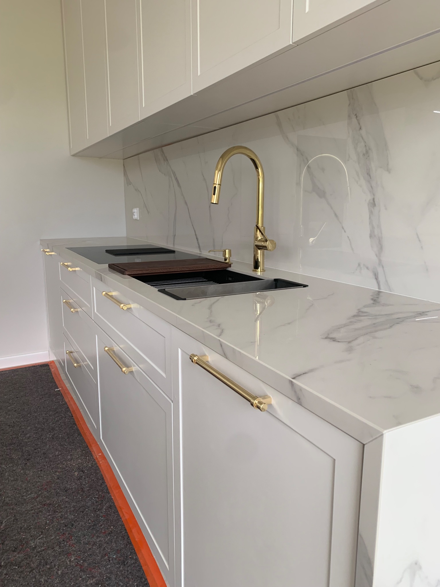 Biała, nowoczesna kuchnia z marmurowym blatem i złotą armaturą. Widoczny zlew, płyta grzewcza i eleganckie uchwyty. Minimalistyczny design.