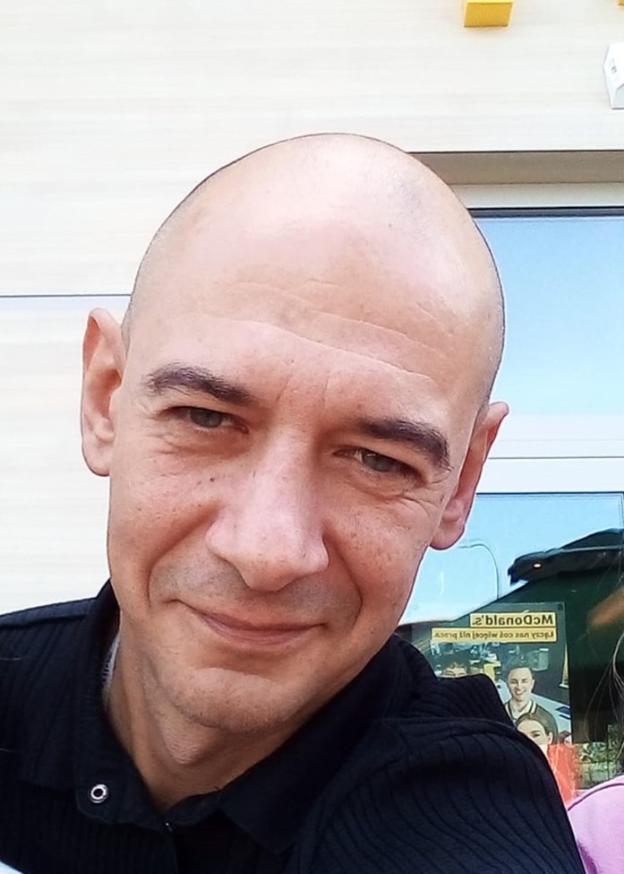 Portret łysiejącego mężczyzny w czarnej koszuli polo, z widocznym odbiciem plakatu McDonald's w tle okna. Uśmiech, bezpośrednie spojrzenie.