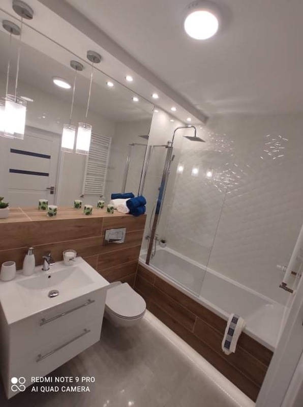 Nowoczesna łazienka z białymi płytkami 3D i drewnopodobnymi akcentami, lustro na całą ścianę, wiszące lampy. Widok na wannę z parawanem i umywalkę z szafką.