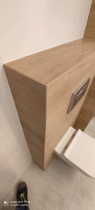 Zabudowa WC w łazience z drewnopodobnych płyt, z widocznym przyciskiem spłukiwania. Ukończony element wykończenia wnętrza łazienki.