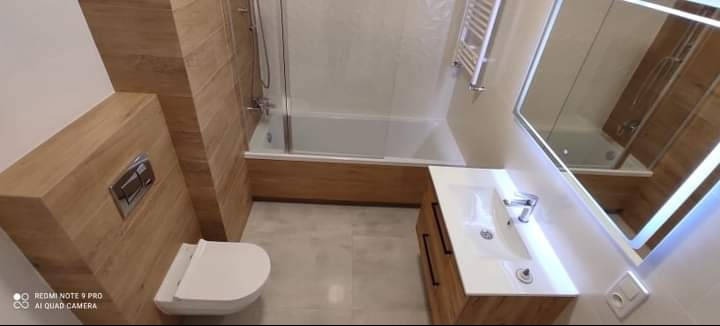 Nowoczesna łazienka z drewnianymi akcentami: umywalka z szafką, toaleta podtynkowa i wanna z parawanem. Lustro z podświetleniem LED.