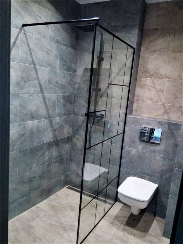 Nowoczesna kabina prysznicowa ze szklaną ścianką w czarnej ramie, szare płytki na ścianach i podłodze, wisząca toaleta. Minimalistyczny design łazienki.