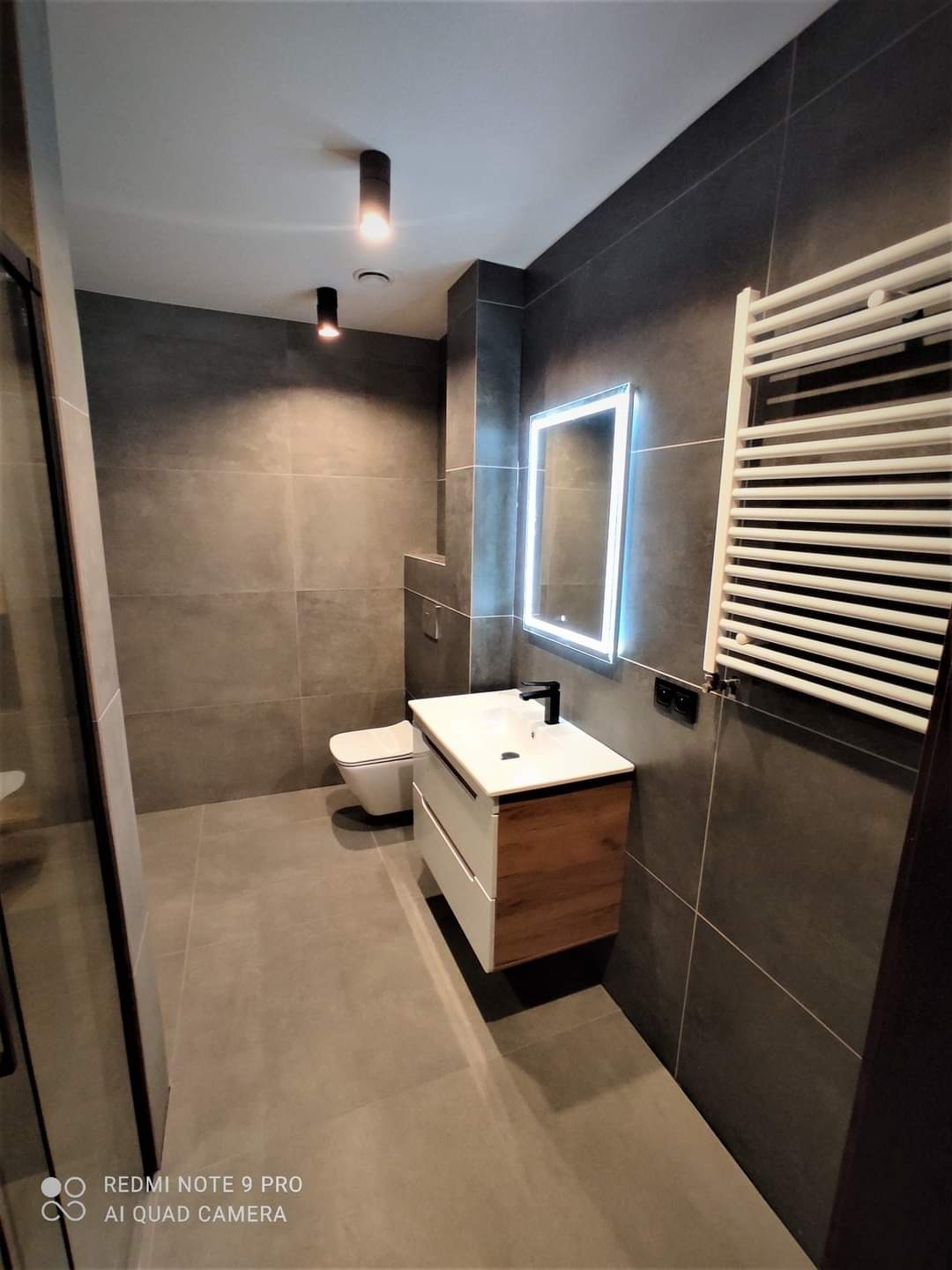 Nowoczesna łazienka w odcieniach szarości z podświetlanym lustrem, wiszącą szafką umywalkową i grzejnikiem drabinkowym. Minimalistyczny design i eleganckie wykończenie.