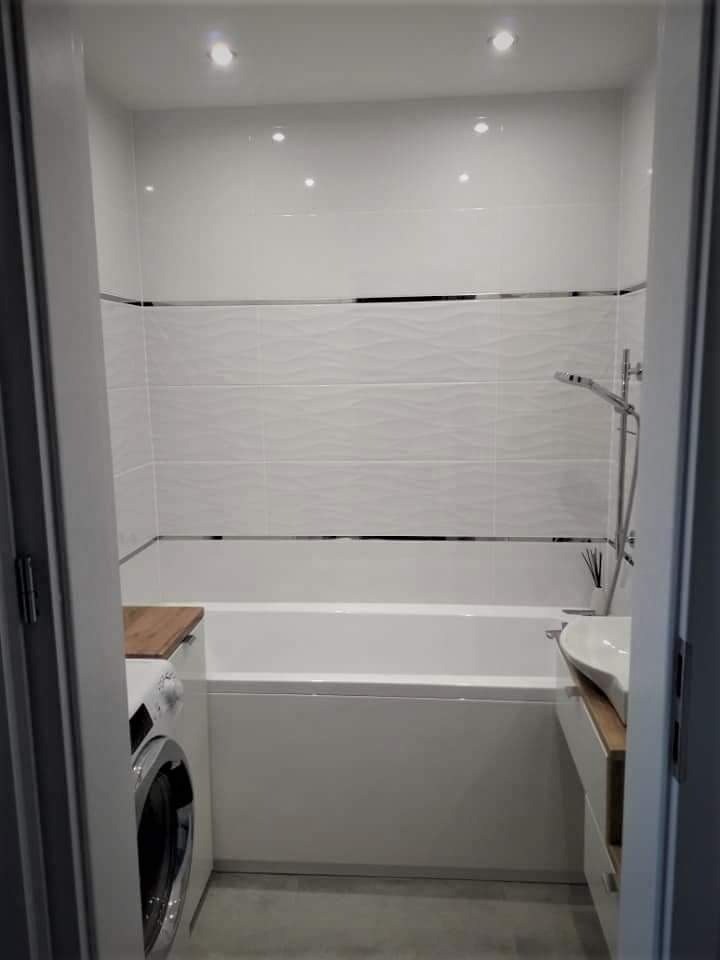 Biała łazienka z wanną, umywalką i pralką w zabudowie. Płytki z subtelnym wzorem fali, oświetlenie punktowe. Minimalistyczny design.