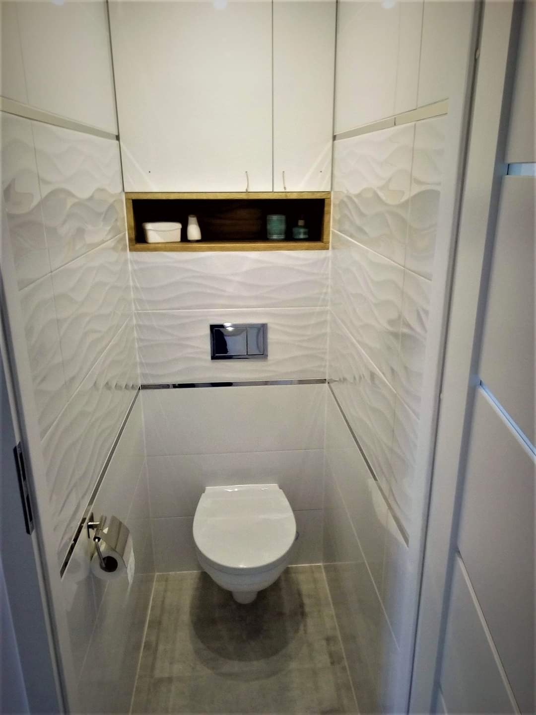 Nowoczesna toaleta w Lubaczowie po remoncie. Białe płytki 3D, podwieszana miska WC, szafki i półka z drewnianym akcentem. Minimalistyczny design, czystość i funkcjonalność.