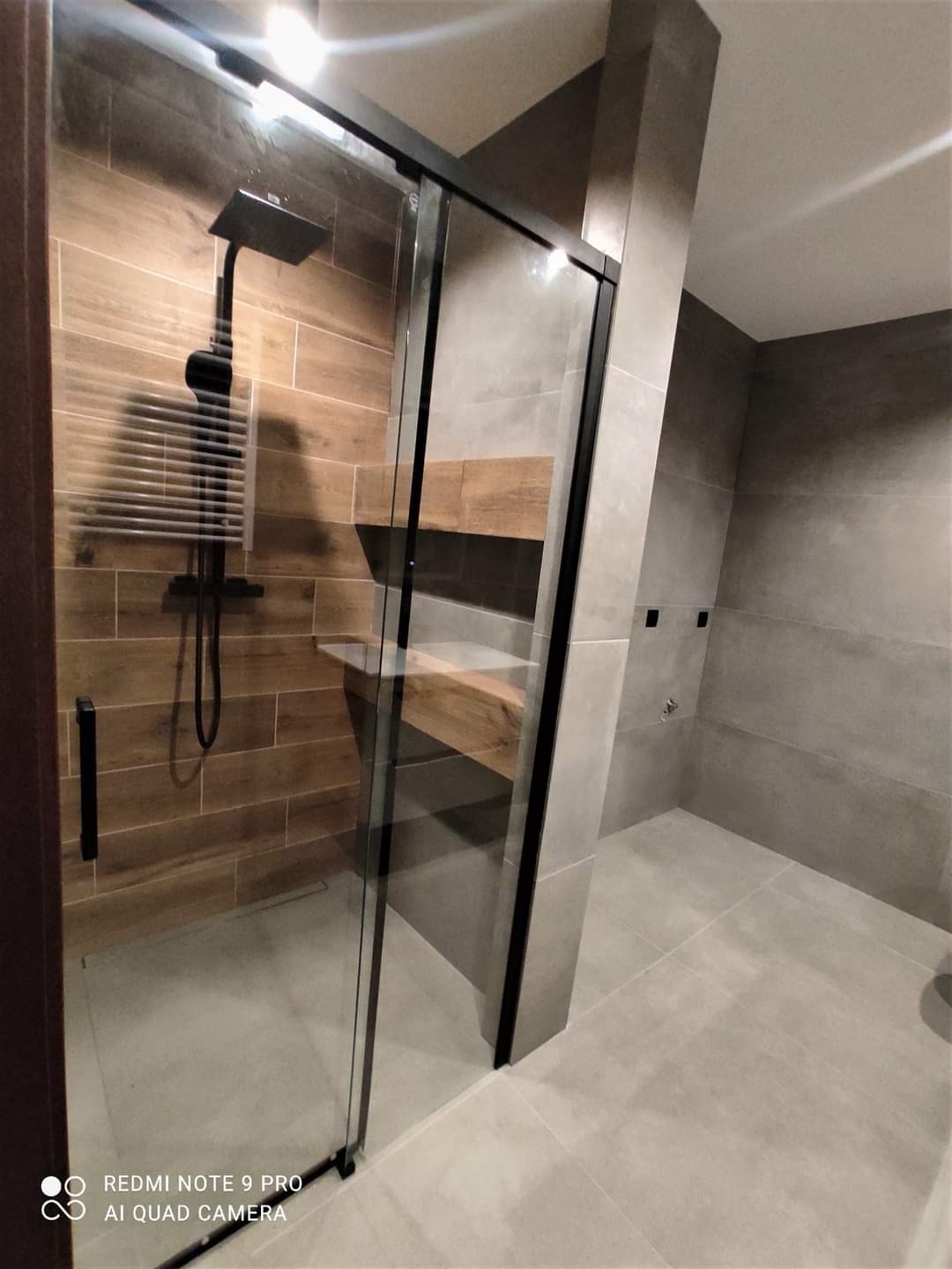 Nowoczesna kabina prysznicowa z czarną ramą, drewnopodobne płytki na ścianie, szare płytki na podłodze i ścianach, minimalistyczny design.