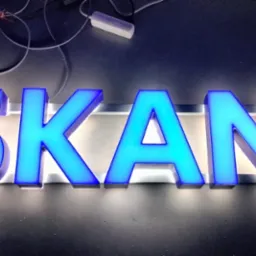 Podświetlany, przestrzenny napis 'SKAN' w odcieniach błękitu i turkusu, umieszczony na ciemnym tle. Widoczne przewody i zasilacz w tle.