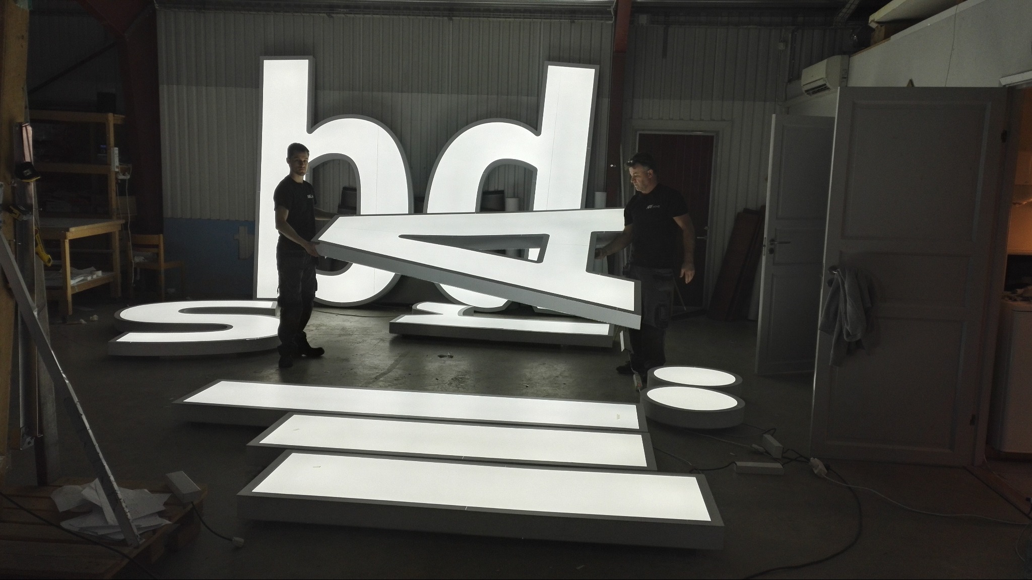 Montaż podświetlanych liter logo 'bd' w warsztacie. Dwóch mężczyzn przenosi element 'A'. Elementy 'S' i 'O' leżą na podłodze. W tle widoczne 'h' i 'd'.