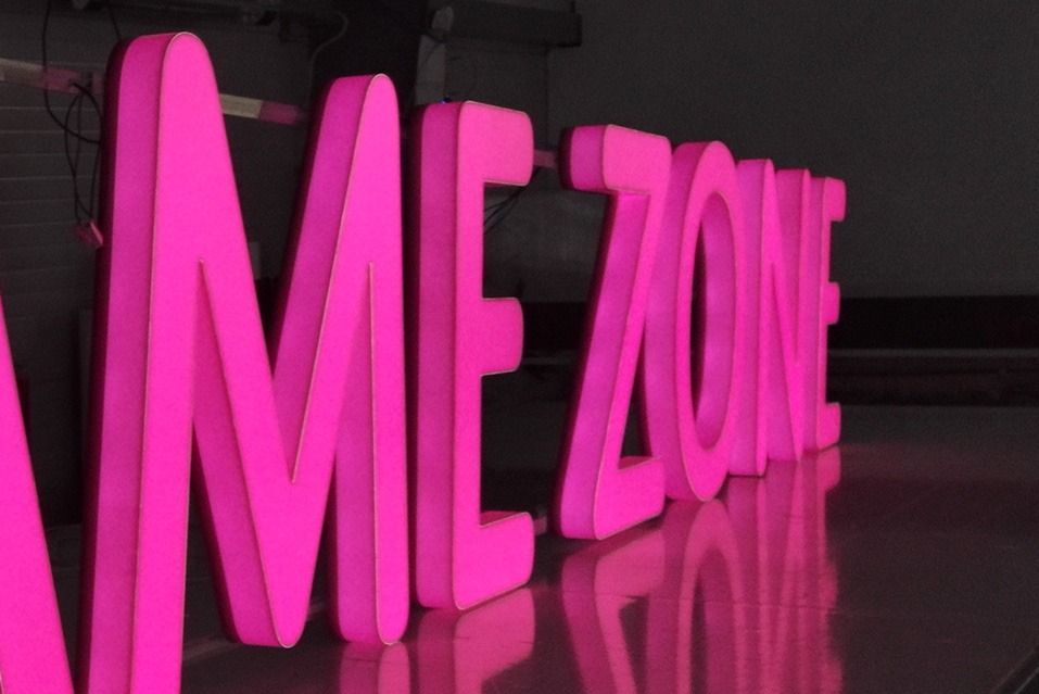 Różowe, podświetlane litery 3D układające się w napis 'ME ZONE', odbijające się w błyszczącej powierzchni, tworząc nowoczesny szyld reklamowy.