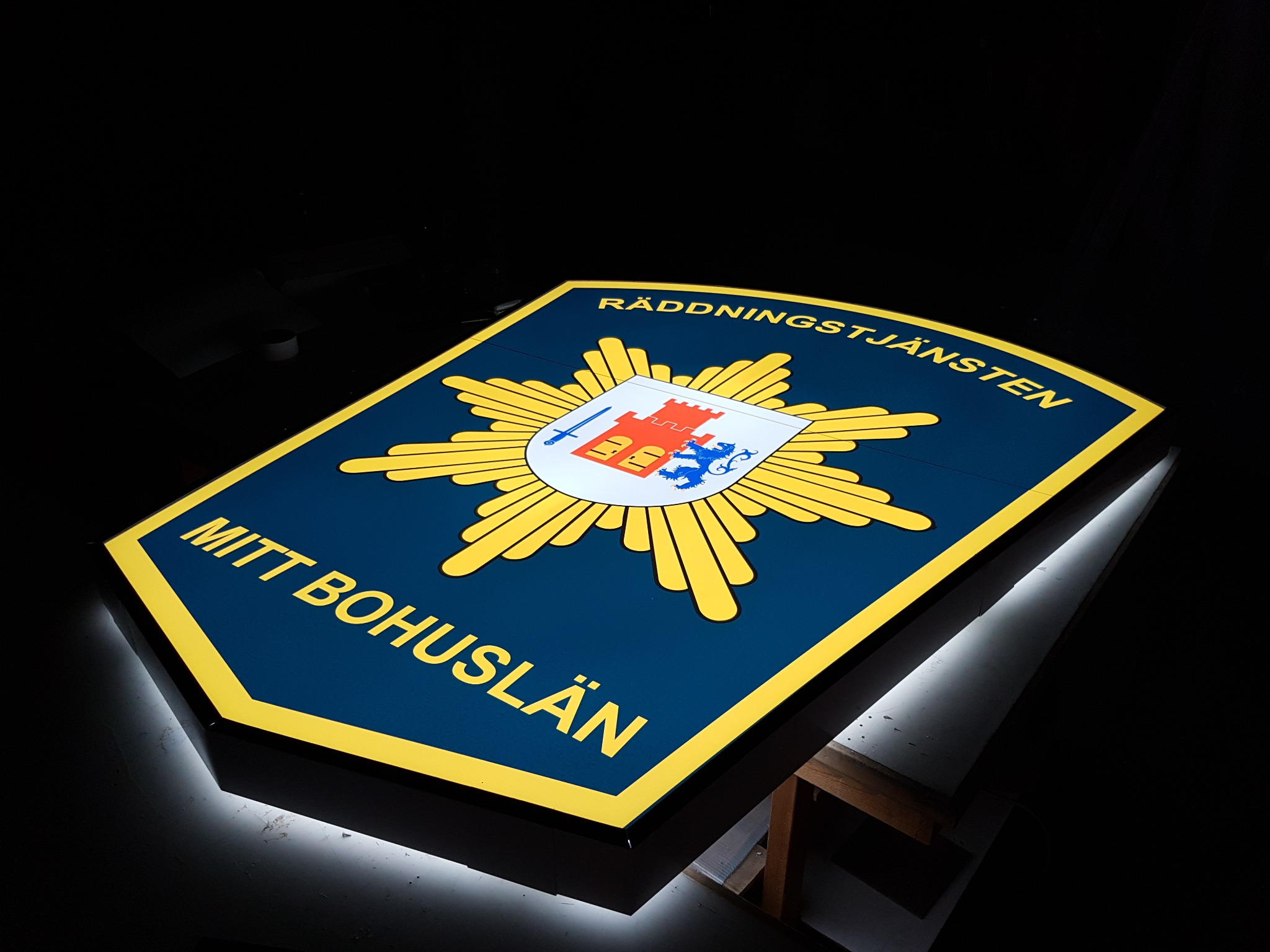 Podświetlany, trójwymiarowy herb RÄDDNINGSTJÄNSTEN MITT BOHUSLÄN w odcieniach niebieskiego, żółtego i białego, z efektem halo na ciemnym tle.