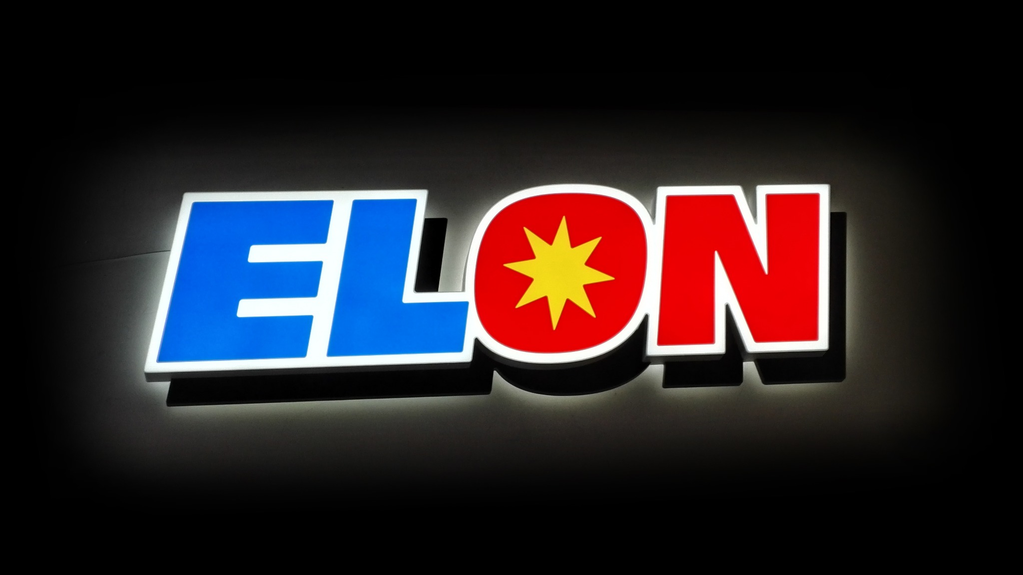 Świecący szyld reklamowy ELON z niebiesko-czerwonym logo na czarnym tle. Litery podświetlone, tworzą efekt 3D.