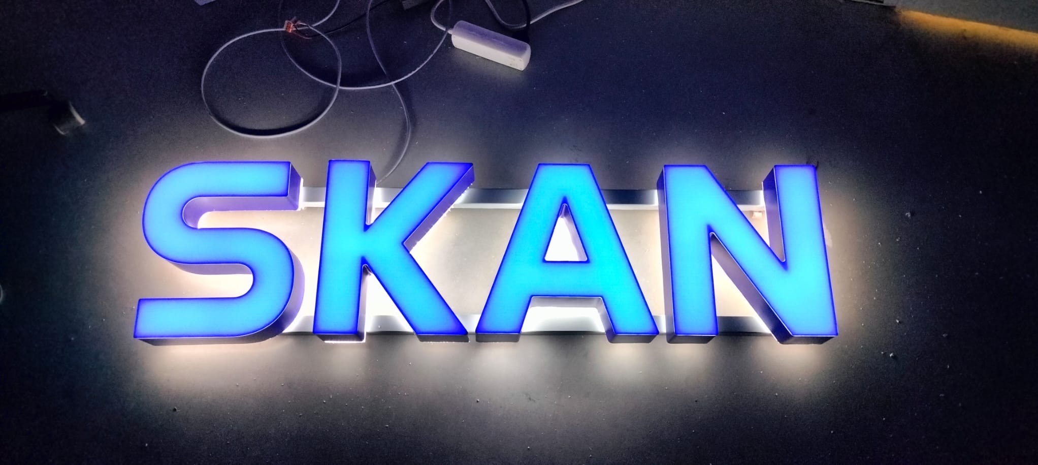 Podświetlany, przestrzenny napis 'SKAN' w odcieniach błękitu i turkusu, umieszczony na ciemnym tle. Widoczne przewody i zasilacz w tle.